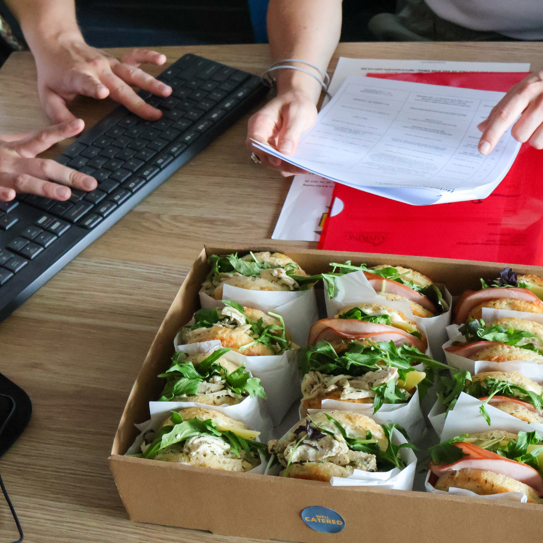 Box of sandwiches on a desk with a person using a computer and holding a document.
