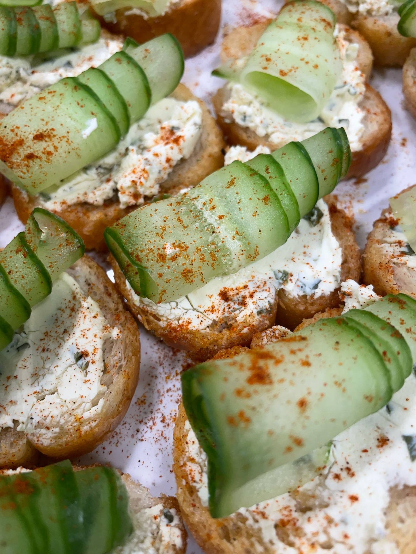Crostini topped with cream cheese, sliced cucumbers, and paprika on a white surface.