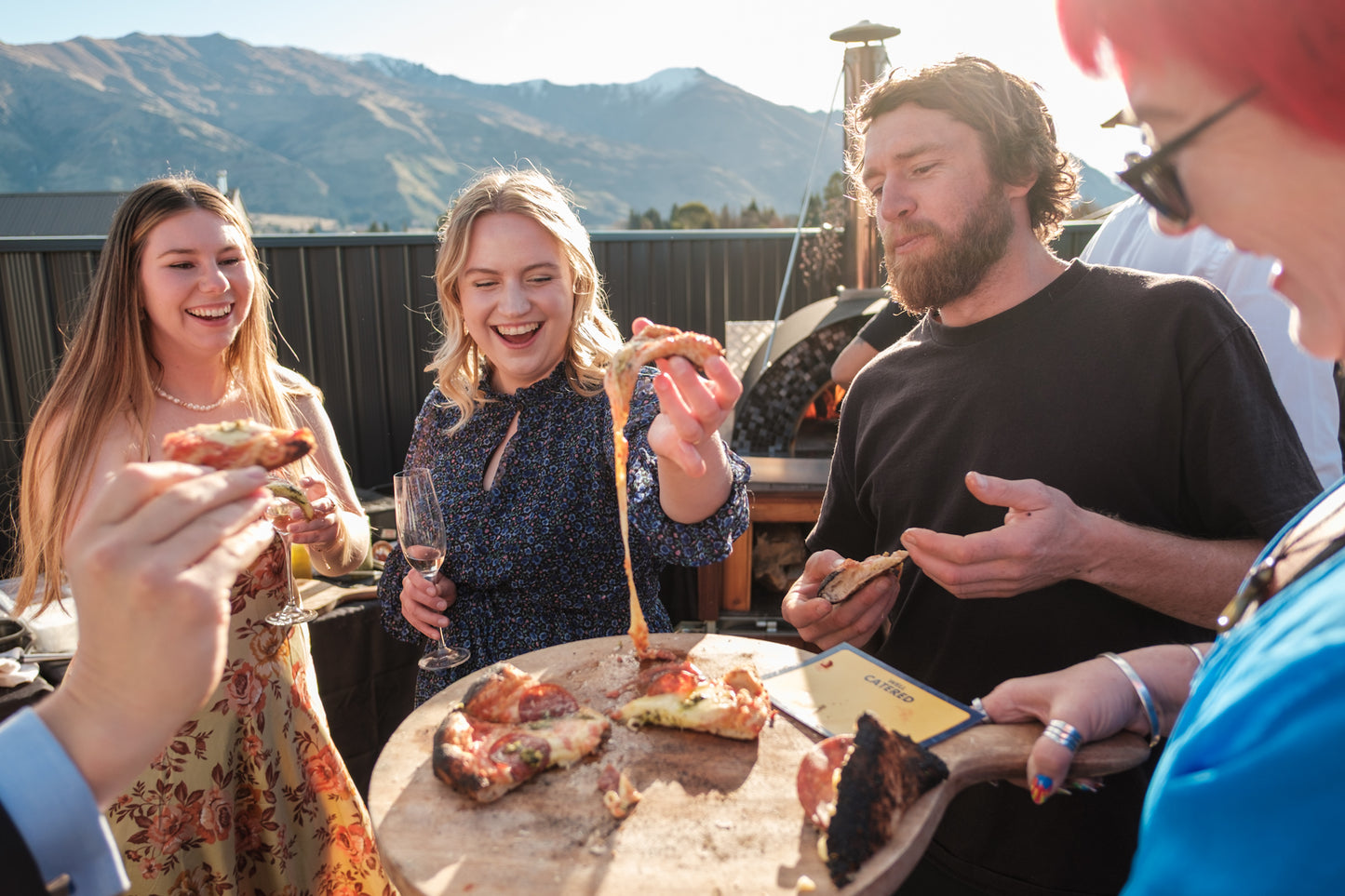 Group of people enjoying pizza on a rooftop with a scenic mountain view.