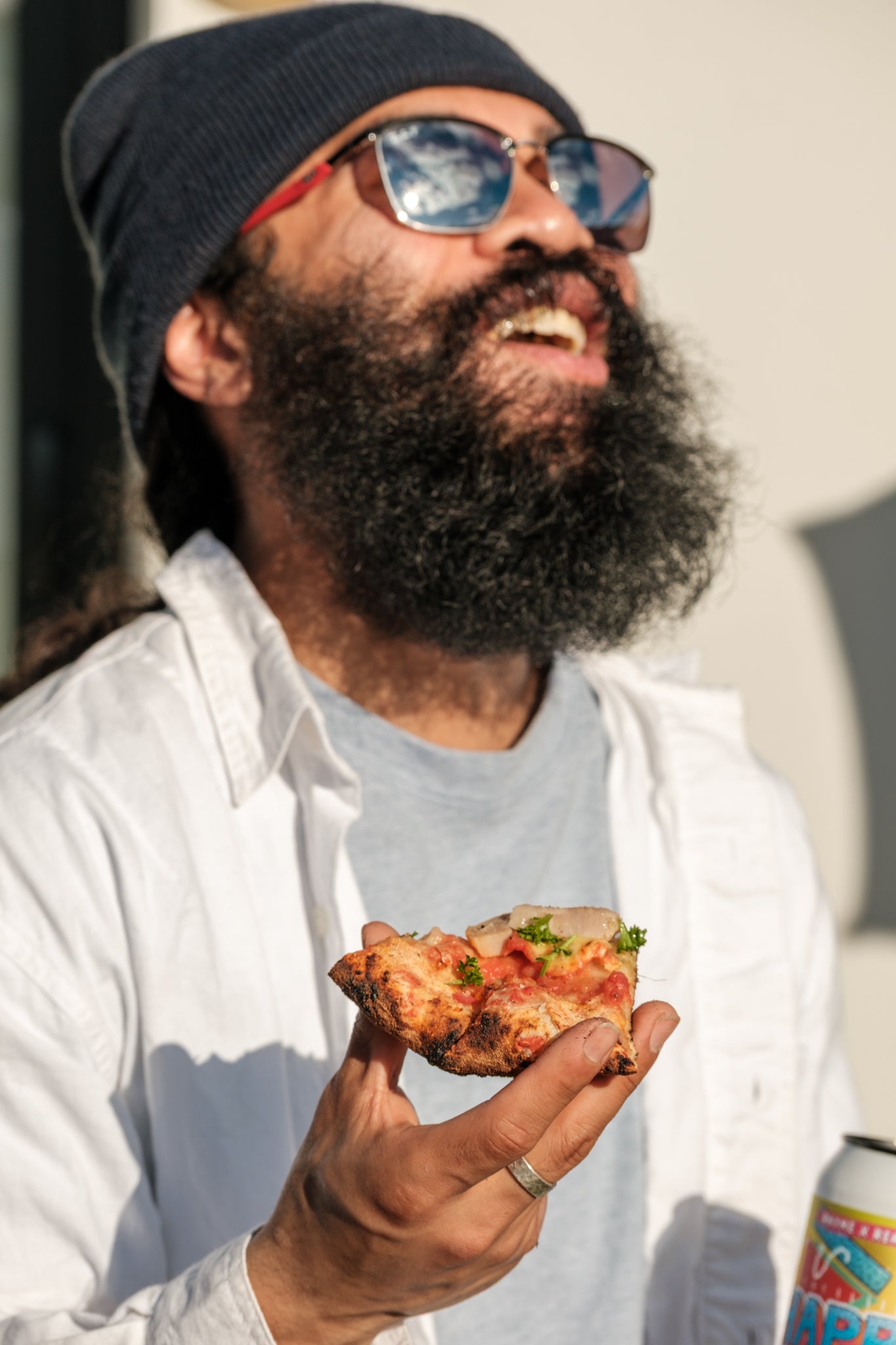 Man with a beard wearing sunglasses and a beanie holding a slice of pizza.
