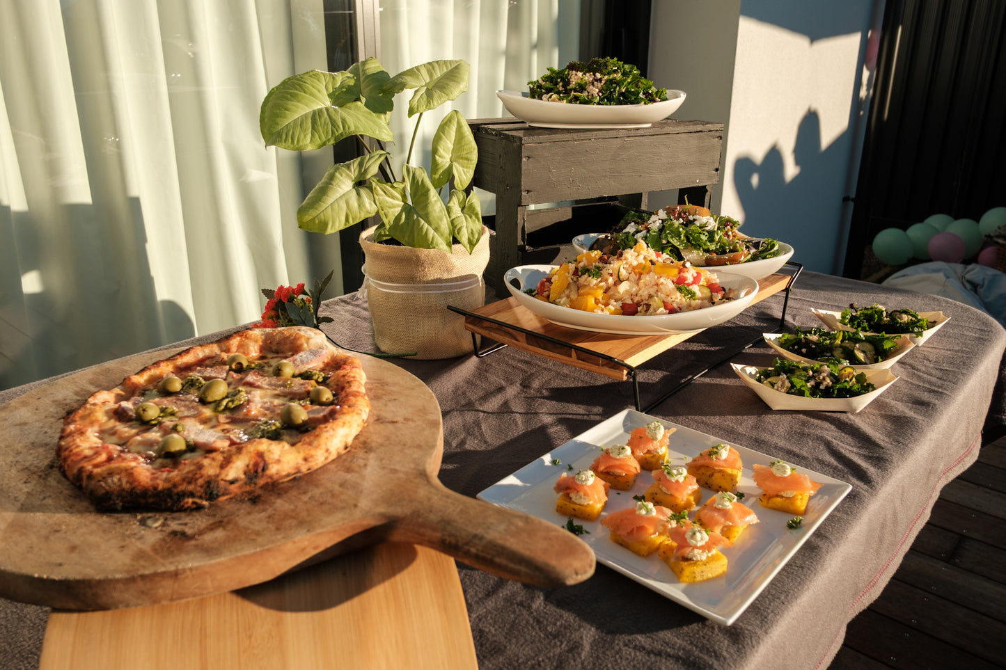 Outdoor setting with a pizza on a wooden board, salads, and a plant on a table.