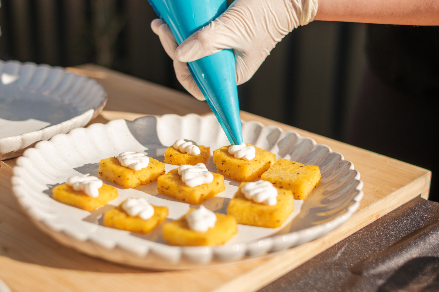 Person decorating small square pastries with white cream using a blue piping bag on a wooden table.