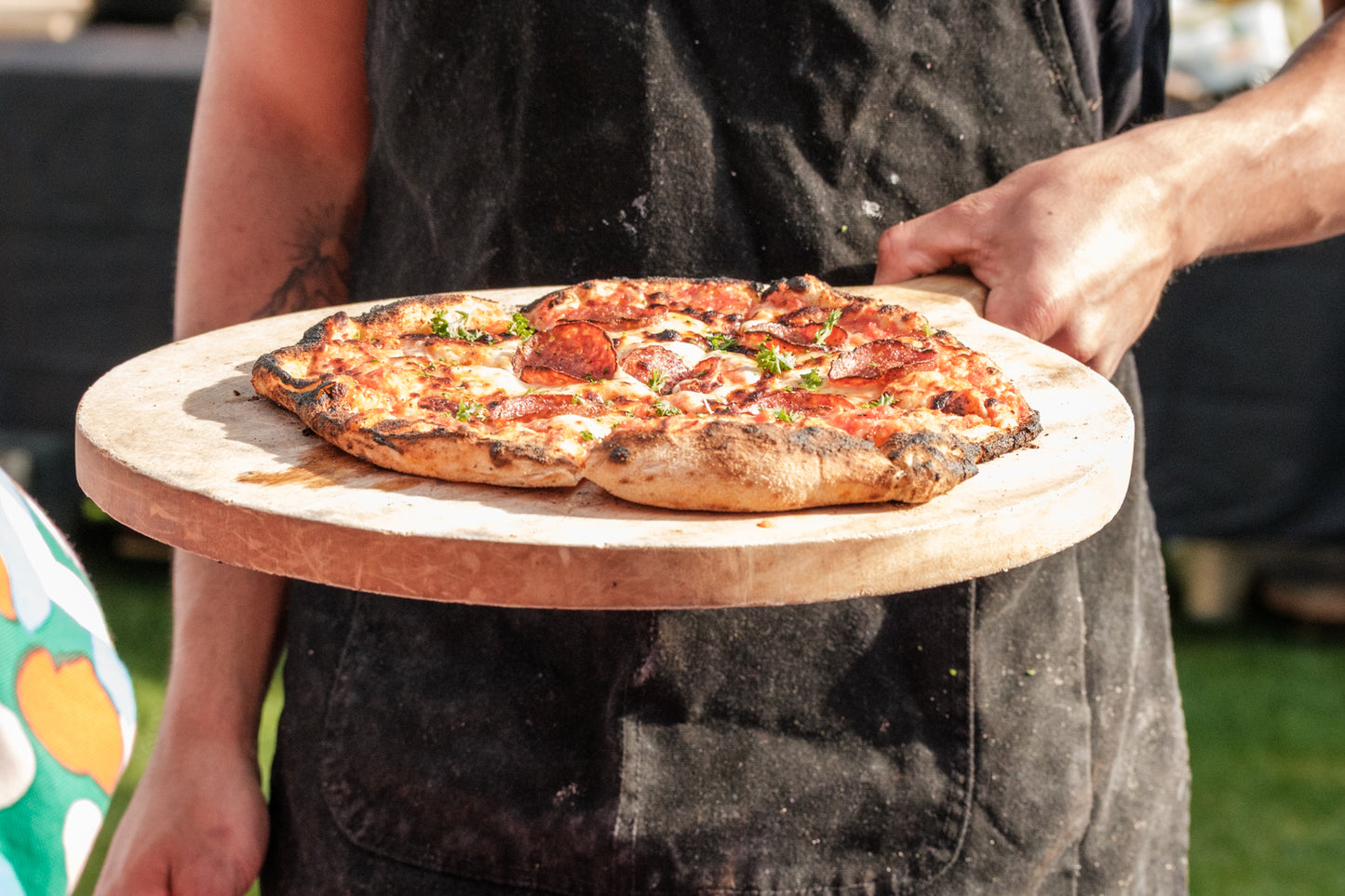Person holding a pizza on a wooden board outdoors