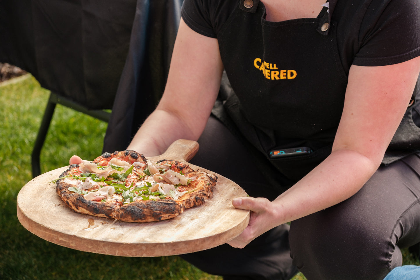 Person holding a pizza on a wooden board outdoors, wearing a black apron with 'Well Catered' branding.