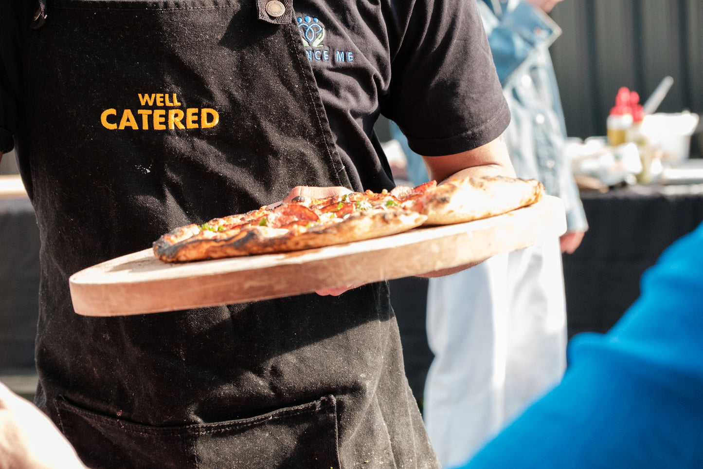Person holding a pizza on a wooden board with 'Well Catered' logo in the background