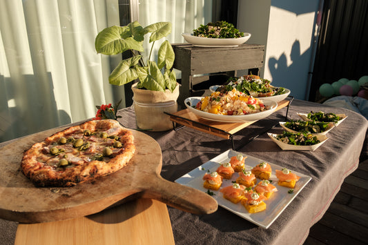Outdoor setting with a pizza on a wooden board, salads, and a plant on a table.