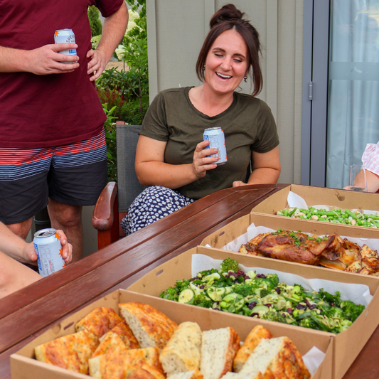 People enjoying food and drinks outdoors, with a focus on a woman holding a can and a table with boxes of food.