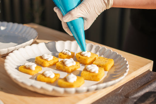 Person decorating small square pastries with white cream using a blue piping bag on a wooden table.