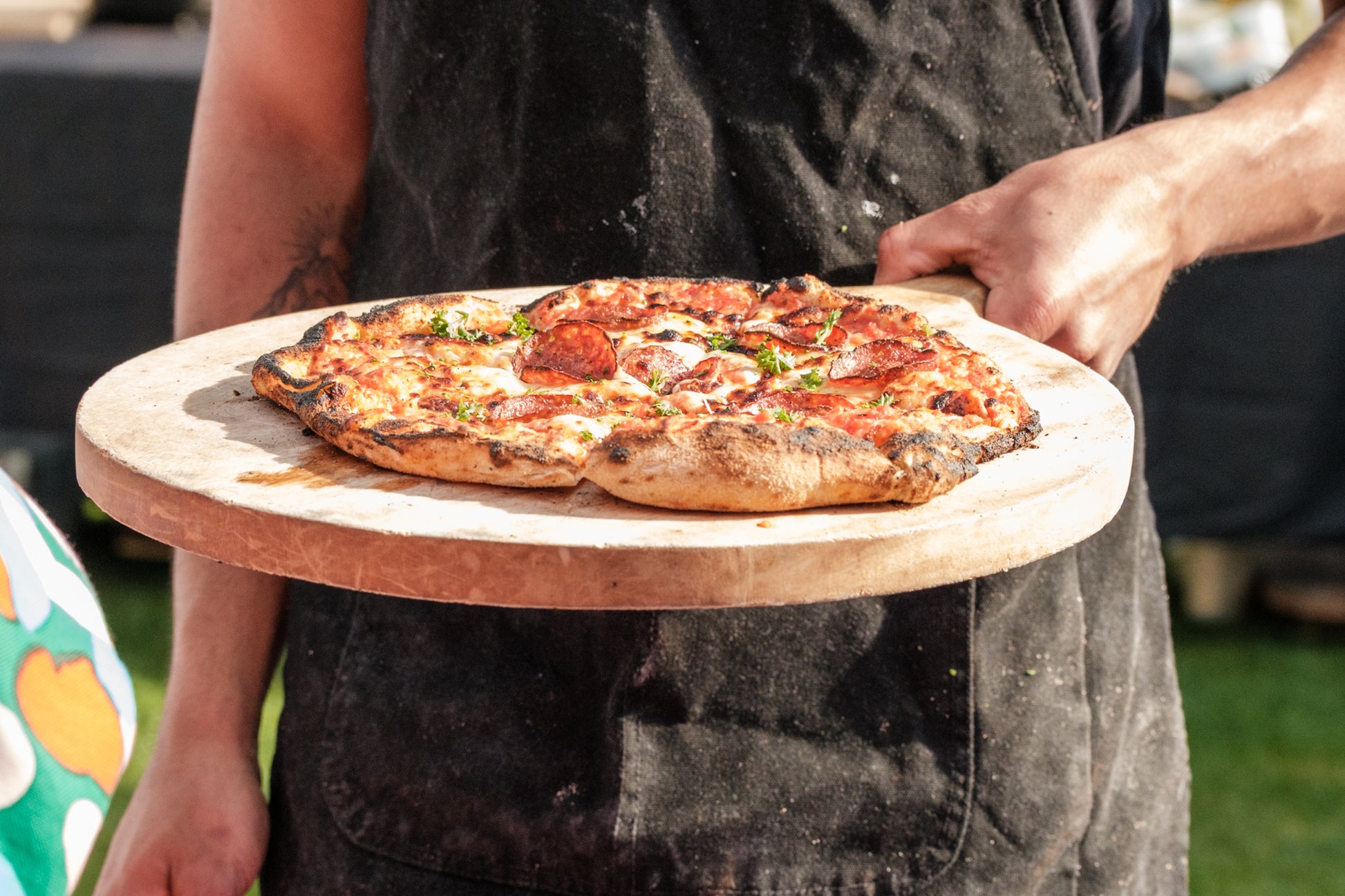 Person holding a pizza on a wooden board outdoors